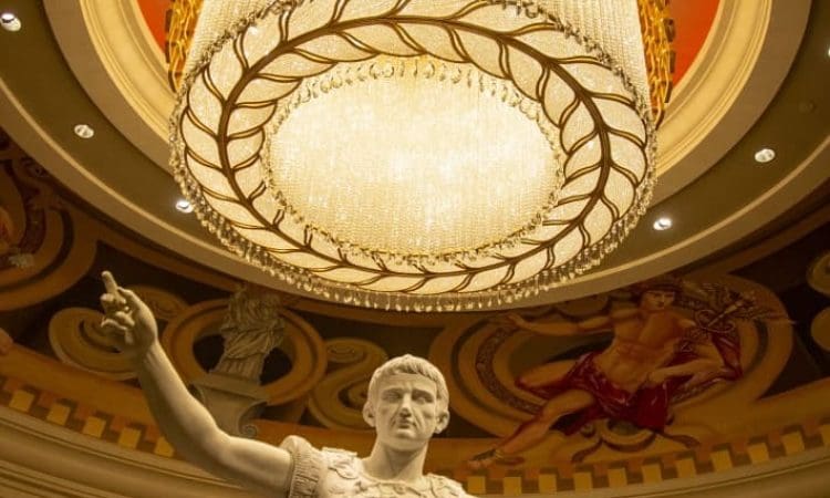 A marble statue of a Roman figure stands beneath a large, ornate chandelier in an elaborately decorated interior with painted ceilings and columns.
