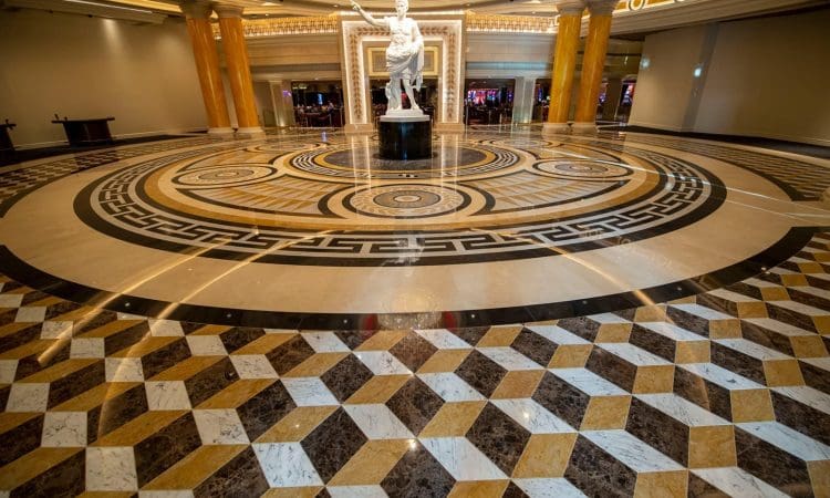 Marble floor with geometric patterns and a statue of a Roman figure in the center of a grand, well-lit hall with gold accents.