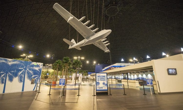 A large model airplane is suspended from the ceiling inside a spacious, dome-roofed exhibition hall with informational signs and palm tree decorations.
