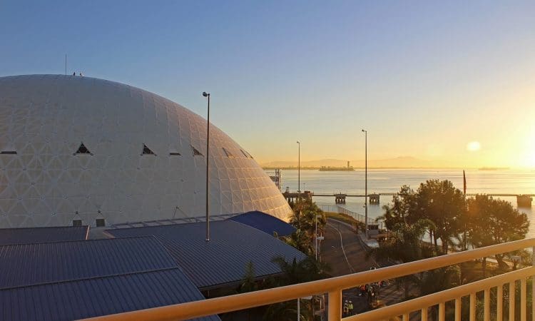 A large white geodesic dome building near a waterfront at sunrise, with a road, trees, and distant cityscape visible.