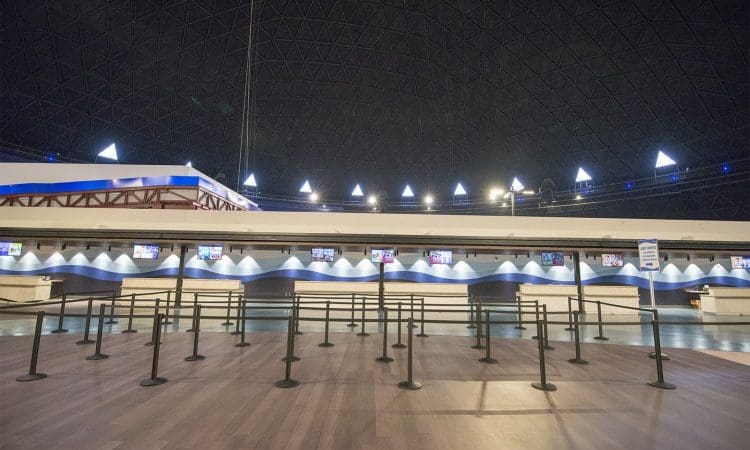 Empty indoor queue area with black stanchions, wooden floor, high domed ceiling, and rows of check-in counters with monitors in the background.