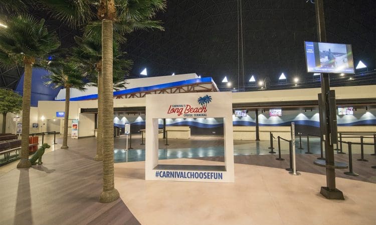 Indoor area of the Carnival Long Beach Cruise Terminal with palm trees, check-in counters, empty queue lines, and a sign reading "#CARNIVALCHOOSEFUN".