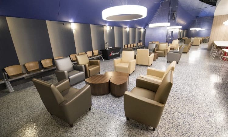 A spacious lounge area with beige and gray armchairs, wooden tables, and modern overhead lighting, featuring a polished speckled floor and a long row of chairs along the wall.