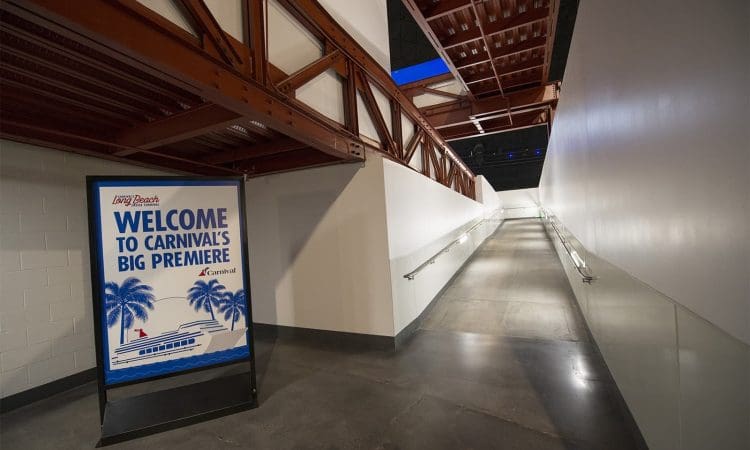 A sign welcoming guests to Carnival’s big premiere stands at the entrance of a long, modern, well-lit hallway with handrails and exposed red beams overhead.