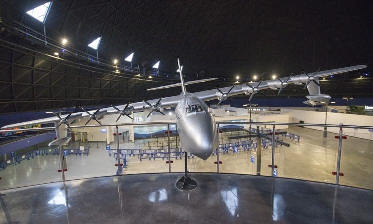 Large seaplane displayed indoors under a domed ceiling, viewed from the front behind a glass barrier, with empty queue lines visible below.