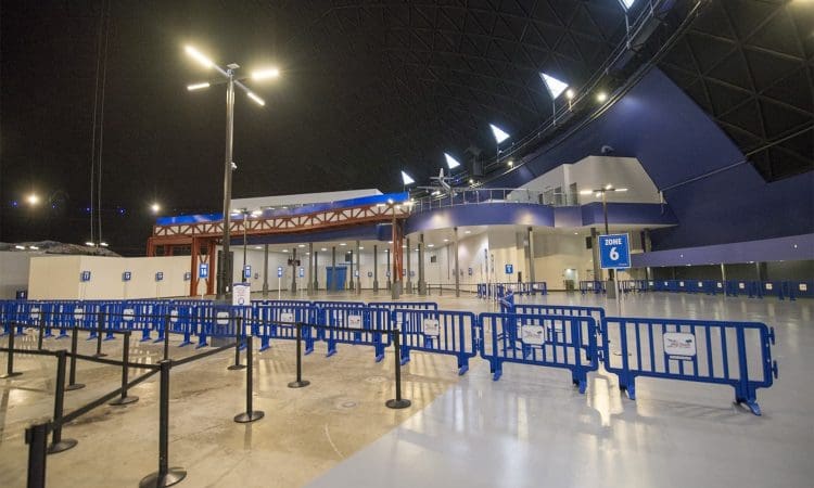 Large indoor venue with blue barriers forming queue lanes, numbered zone signs, and bright overhead lights. The space appears empty and clean.