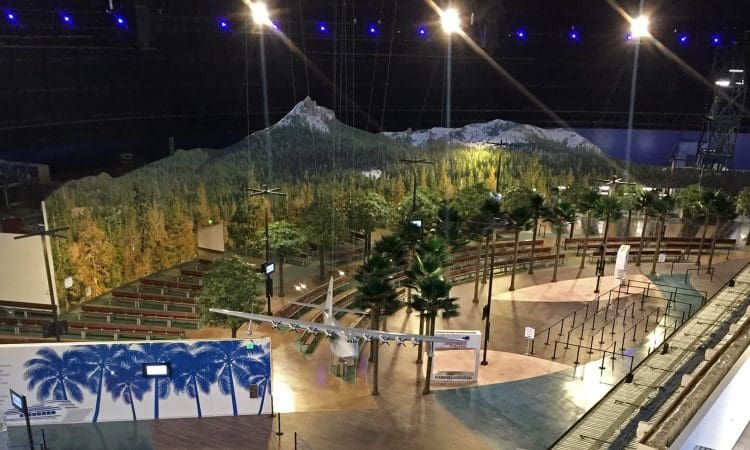 Indoor area with artificial palm trees, benches, and a large mountain forest mural in the background; overhead lights and barriers visible.