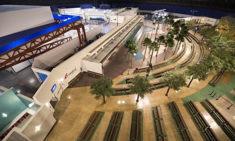A spacious indoor area with benches, artificial trees, curved walkways, and building facades, resembling an airport or transportation hub.