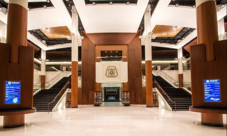 Spacious, modern lobby with high ceilings, wood paneling, two large staircases, digital information screens, and an emblem displayed above the central entrance.