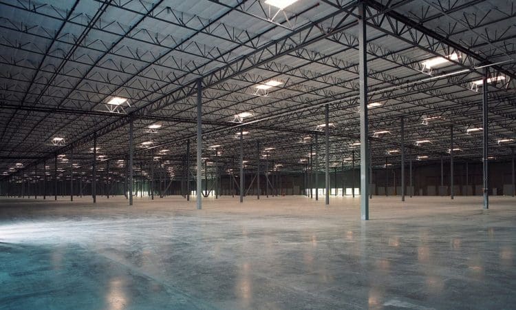 Large, empty warehouse interior with high ceilings, metal support beams, and polished concrete floors, illuminated by overhead lights.