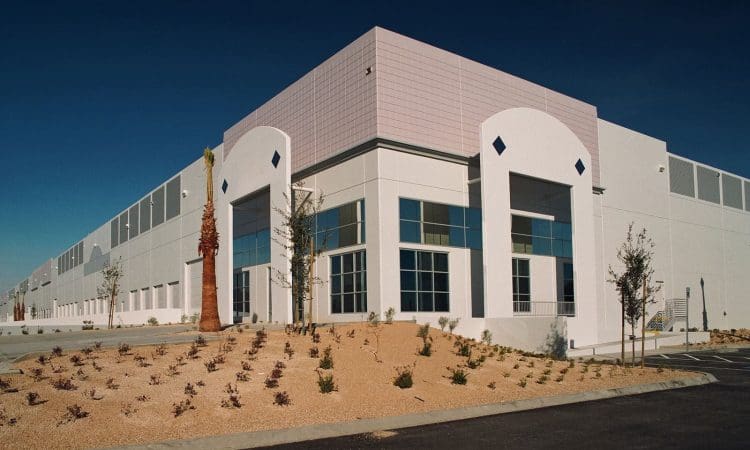 A large, modern industrial building with white exterior walls, large windows, minimal landscaping, and empty parking spaces under a clear sky.