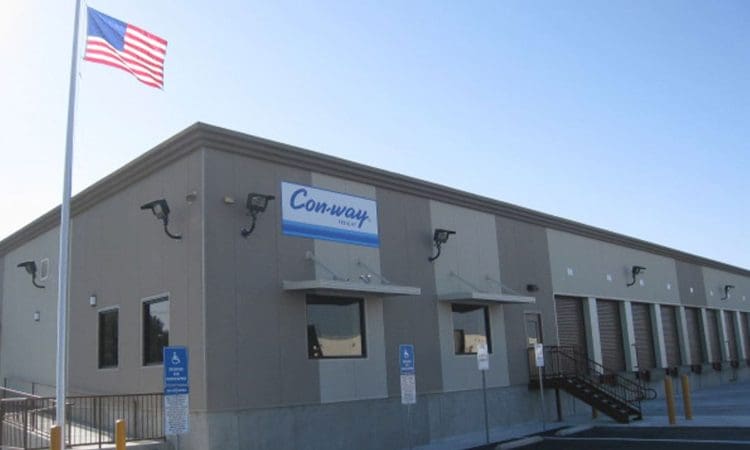 A beige industrial building with a "Con-way Freight" sign, an American flag, multiple loading docks, and designated accessible parking spaces in front.
