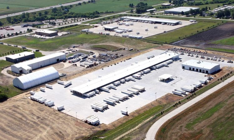 Aerial view of a large industrial facility with multiple warehouses, parked trucks, and open storage areas, surrounded by green fields and nearby buildings.