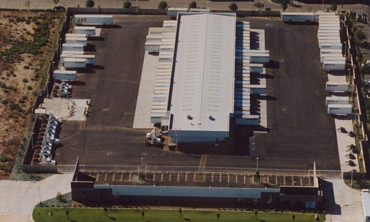 Aerial view of a fenced warehouse facility with multiple truck trailers parked around the building and an adjacent parking lot in front.