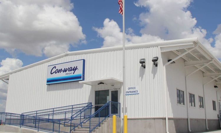 A white industrial building with a "Conway Freight" sign, blue railings, an American flag, and a sign indicating the office entrance. The sky is partly cloudy.