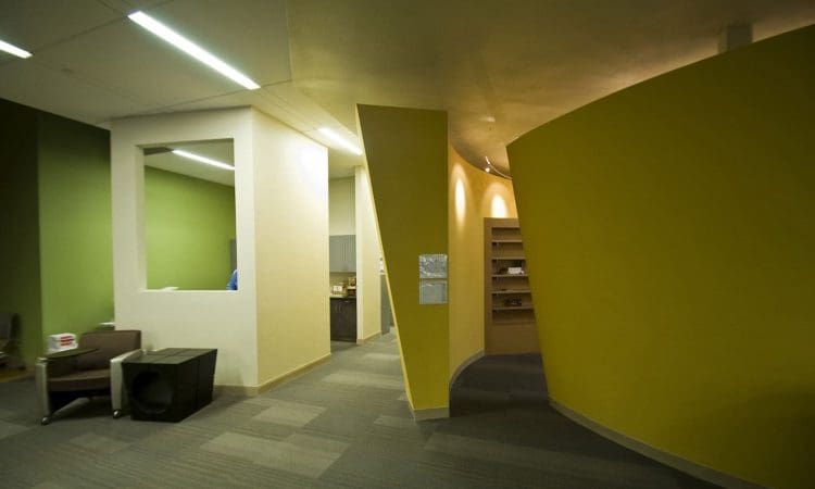 Modern office interior with green and yellow curving walls, a gray armchair, small table, and recessed lighting, featuring a mix of open and enclosed spaces.
