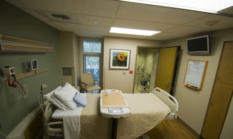 A hospital room with a neatly made bed, medical equipment on the wall, a TV, a whiteboard, a chair by the window, and a framed flower picture on the wall.