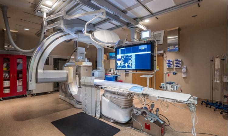 Modern hospital angiography suite with advanced imaging equipment, monitors displaying medical scans, and various medical devices arranged around the patient table.