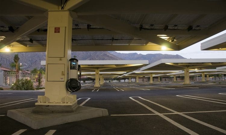A mostly empty parking lot with solar panel canopies and an electric vehicle charging station attached to a support column. Mountains are visible in the background.