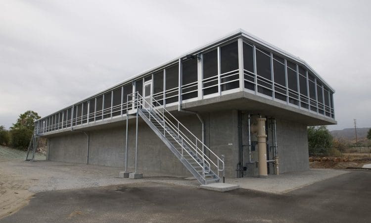 A modern, elevated industrial building with metal railings and an exterior staircase, set on a gravel lot under an overcast sky.