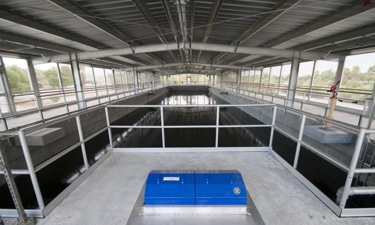 Interior view of a modern water treatment facility featuring a large rectangular tank, metal railings, and a blue equipment box in the foreground under a metal roof.