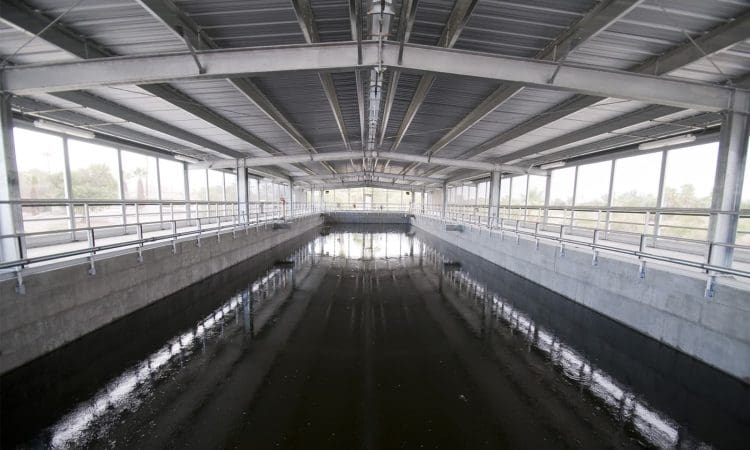 A large, covered water treatment facility with a concrete basin and metal roof, featuring railings along both sides and water pooled at the bottom.