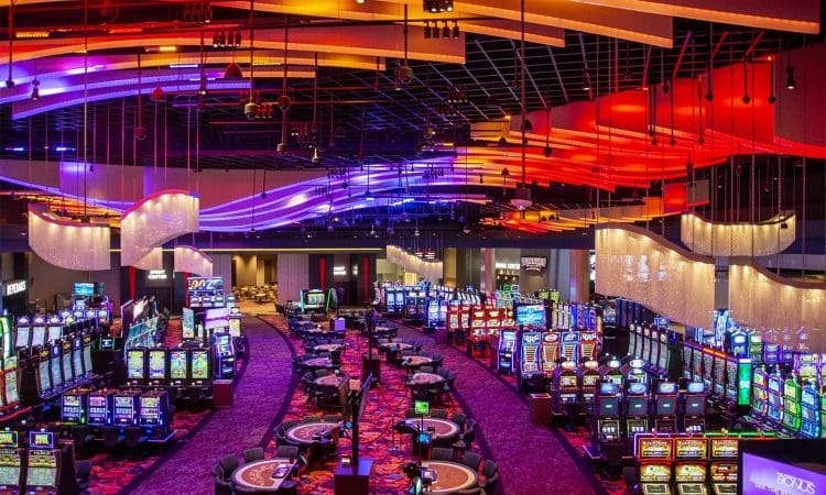 Casino interior with rows of slot machines, gaming tables, and colorful ceiling lights in purple and red hues.