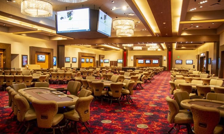 Spacious casino poker room with multiple card tables, empty chairs, red patterned carpet, large chandeliers, and several wall-mounted screens.