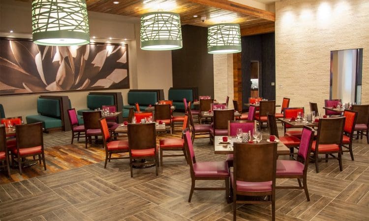 Modern restaurant interior with colorful chairs around wooden tables, green booth seating, large hanging lights, and a large flower artwork on the wall.