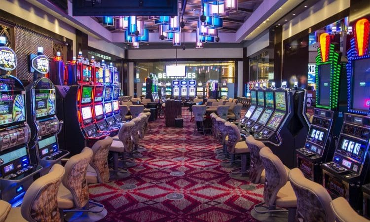 Casino interior with rows of empty slot machines on both sides, patterned carpet, and modern ceiling lights, creating a clean and organized gaming atmosphere.