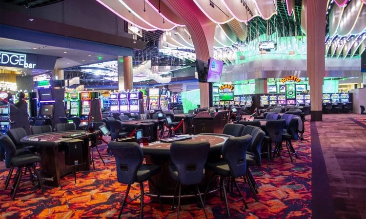 Empty casino gaming floor with rows of slot machines, gaming tables, and colorful carpeting under bright lights.