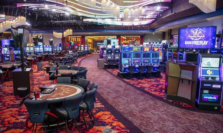 Casino interior with rows of slot machines, electronic gaming stations, and card tables under colorful ceiling lights. The space is brightly lit and mostly unoccupied.