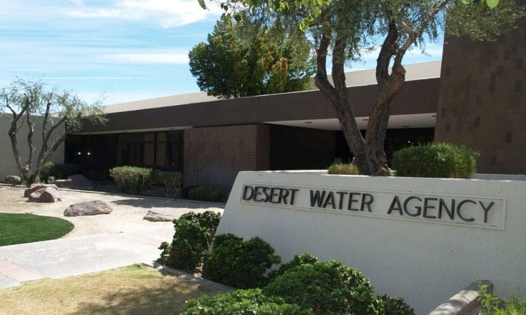The exterior of the Desert Water Agency building, featuring a sign in front, trees, shrubs, and a clear sky in the background.