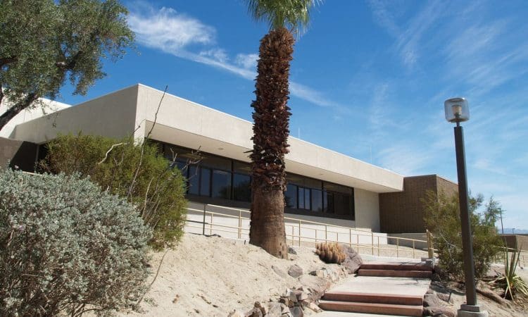 A modern, single-story building with large windows, surrounded by desert plants and a palm tree, under a blue sky with scattered clouds.