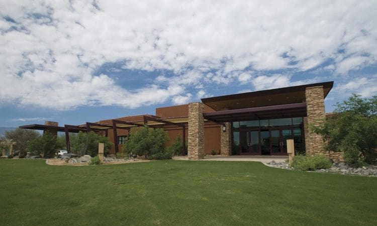 Modern building with stone pillars and large glass windows, surrounded by green lawn and desert landscaping under a partly cloudy sky.