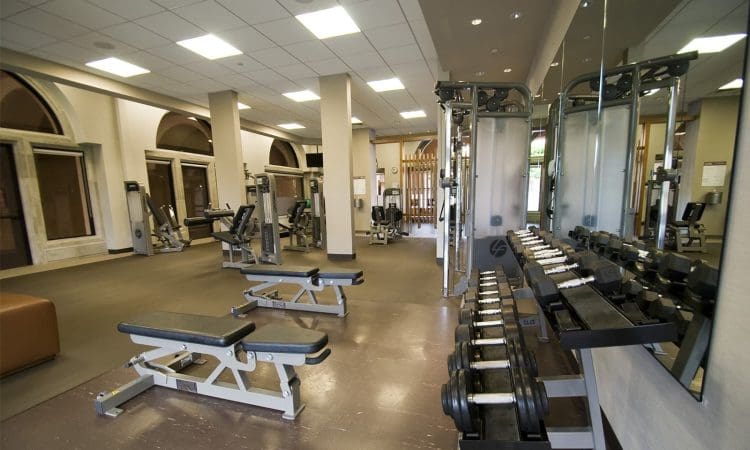 A fitness gym with benches, weight racks, dumbbells, mirrors, and various exercise machines in a well-lit room with high ceilings.