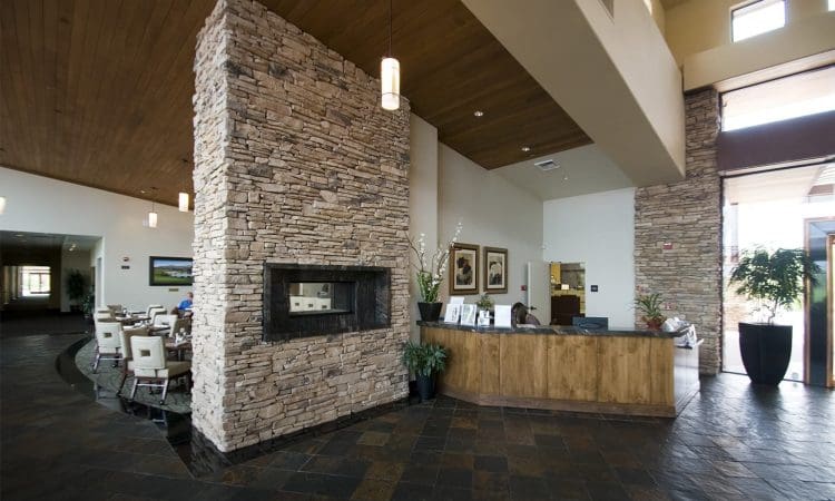 A modern lobby with a stone fireplace, wooden reception desk, tile flooring, high ceilings, and dining area visible in the background.