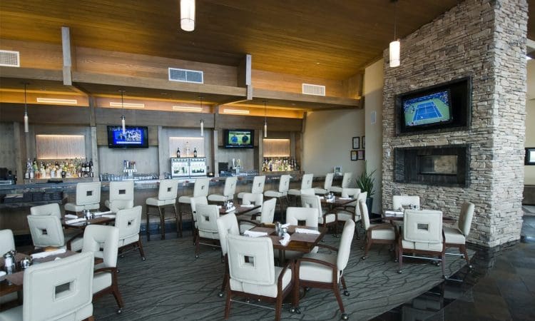 Modern restaurant bar with white cushioned chairs, set tables, a stone fireplace, and several TVs on the walls displaying different channels.
