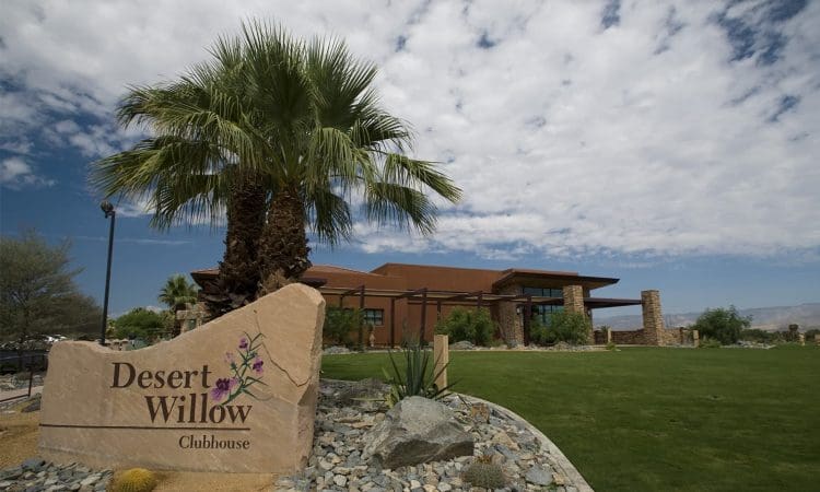 A clubhouse building sits behind a landscaped area with palm trees and a large stone sign reading "Desert Willow Clubhouse" under a partly cloudy sky.