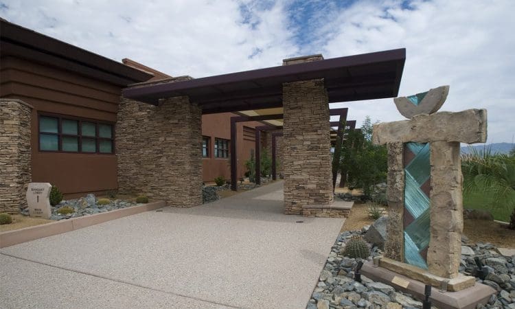 A modern building with stone columns, a covered walkway, and a geometric sculpture in a landscaped area with rocks and desert plants.