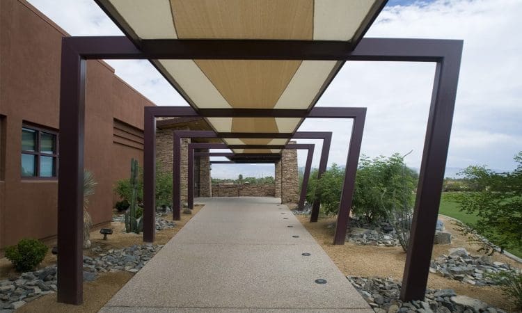 Covered outdoor walkway with metal frame and beige fabric canopy, bordered by rocks, plants, and a red building on the left.