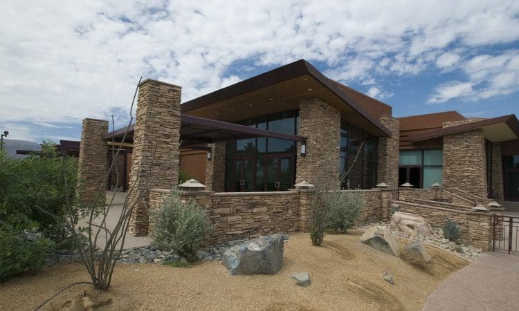 Modern building with stone columns and walls, large glass windows, and a landscaped area featuring desert plants and rocks under a partly cloudy sky.