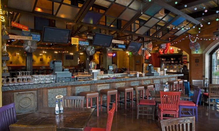 A bar and restaurant interior with colorful wooden chairs, a long counter lined with barstools, hanging TVs, and decorative signs and lights.