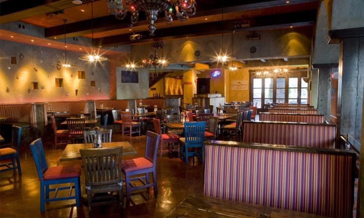 Spacious restaurant interior with wooden tables, colorful chairs, striped booth seating, and decorative lighting fixtures. The space is empty of people.