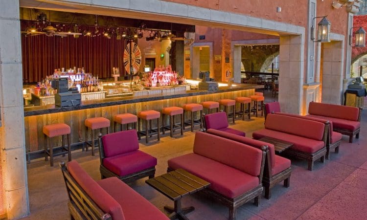 Indoor bar with a long counter, red bar stools, and red cushioned seating areas; bottles and glasses are visible behind the bar, with stage and lighting in the background.
