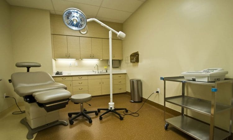 A medical examination room with an exam chair, overhead light, stools, a metal cart, sink, cabinets, and medical equipment.