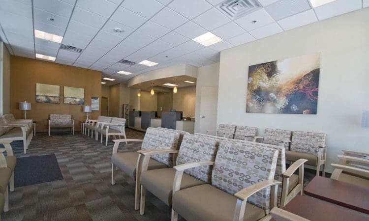 A modern waiting room with patterned chairs, neutral-colored walls, abstract artwork, and a reception desk in the background. The space is empty.