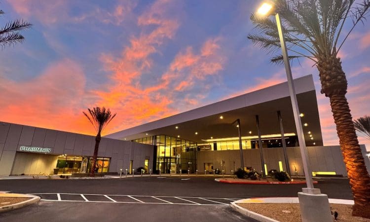 Modern building with large glass windows and a pharmacy sign, illuminated at dusk with palm trees and a vibrant sunset sky in the background.