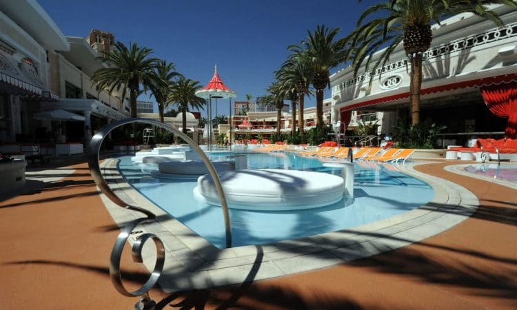 Outdoor swimming pool area with palm trees, lounge chairs, and round in-pool seating surrounded by buildings under a clear blue sky.