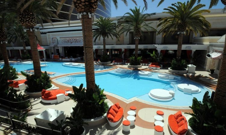 Luxurious outdoor pool area with palm trees, red and white lounge chairs, circular tables, and modern decor under a clear blue sky.